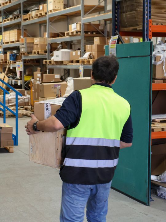 Warehouse worker in a hi-vis vest crouching between shelving aisles stocked with boxed drive and transmission components.