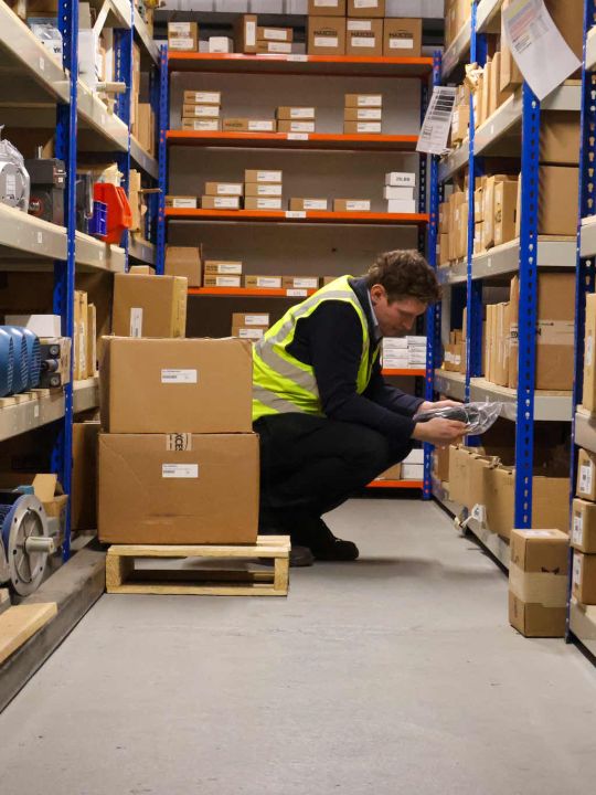 Warehouse worker in a hi-vis vest crouching between shelving aisles stocked with boxed drive and transmission components.