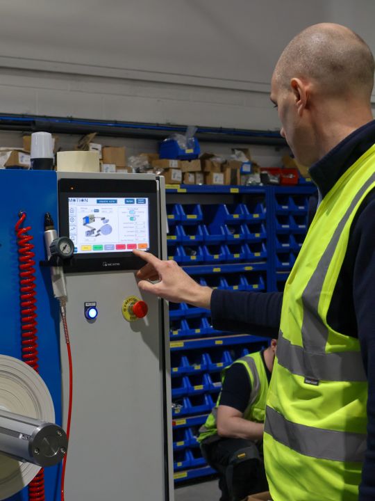 A technician in a hi-vis vest operating a Weintek touchscreen control panel on a Motion Drives and Controls machine in a warehouse.